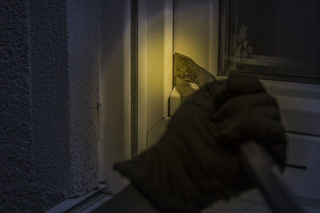 ręka włamywacza w czarnej rękawiczce oświetlona latarką otwiera łomem okno do domu chcąc się włamać i zakłócić spokój i bezpieczeństwo