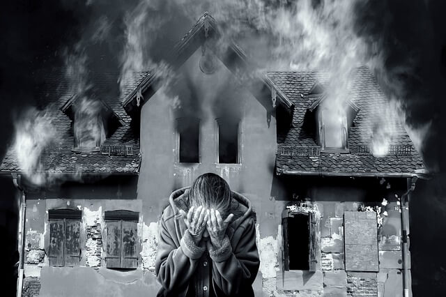 szare zdjęcie kobiety zakrywającej dłońmi twarz na tle palącego się domu który nie był wyposażony w inteligentny czujnik dymu